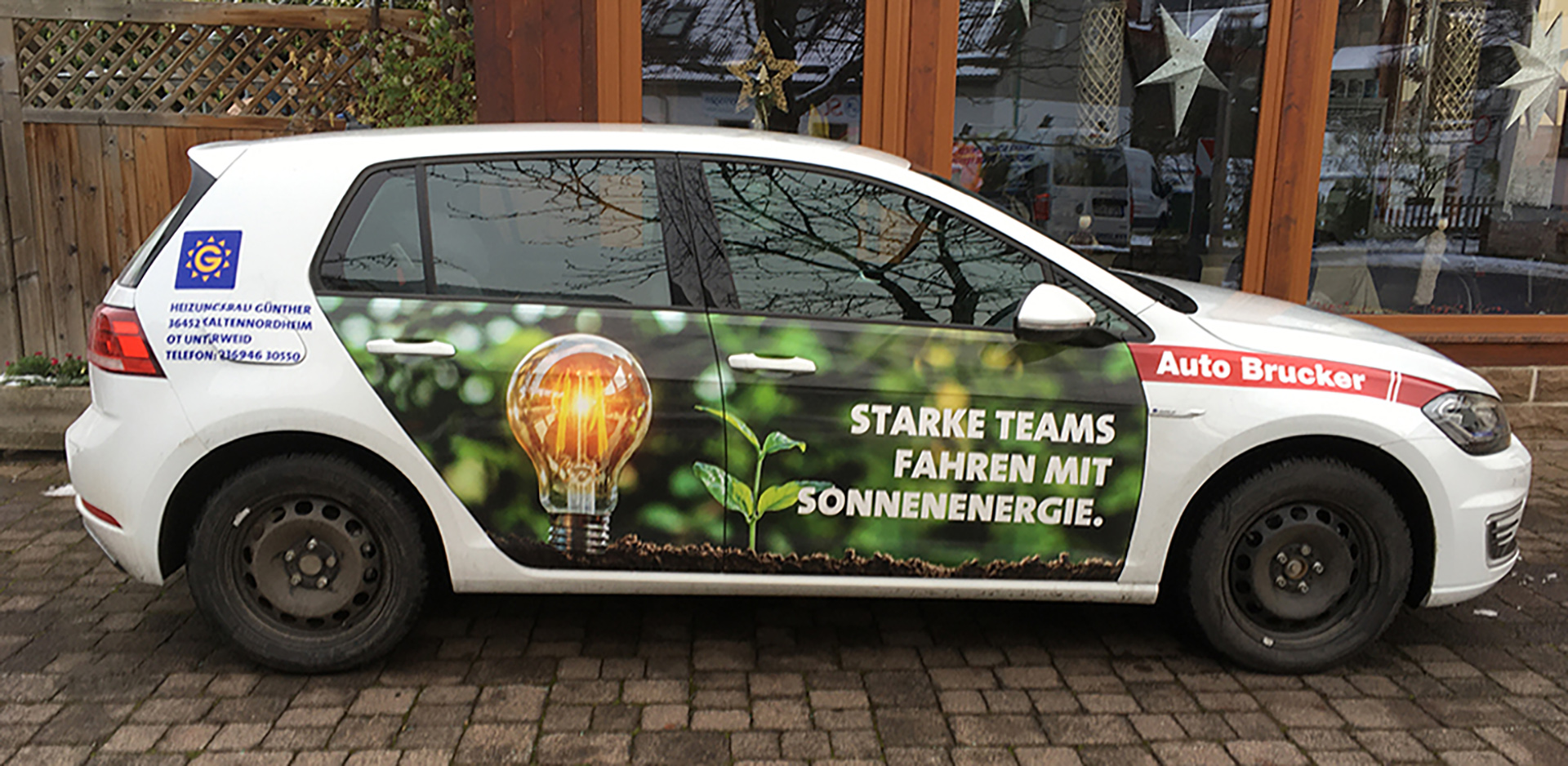 Ein weißes E-Fahrzeug mit einem Bild auf den Türen. Das Bild zeigt eine Glühbirne und die Aufschrift "Starke Teams fahren mit Sonnenenergie."